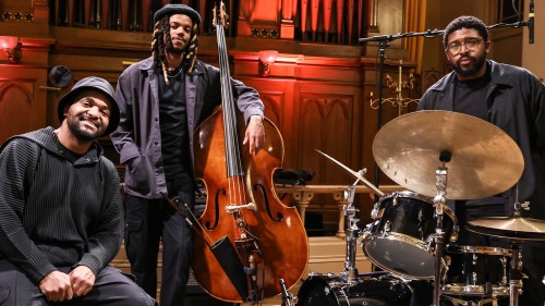 Three black men in black attire stand in front of organ pipes, one sits on a piano bench, the others pose with a bass and drum set. 