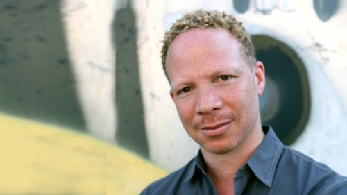 Craig Taborn, a man with light hair stands in front of a patterned wall in a blue shirt smiling. 
