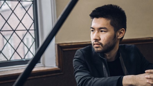 An Asian American man in a black jacket sits leaning on a grand piano, his face framed by the lid and the stick in a well-lit room. 