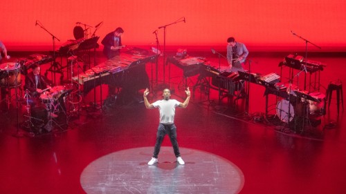 On a stage bathed in red, a single dancer stands in the center with his arms stretched upward, surrounded by mic'd percussion. 