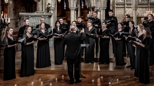 A choir of twenty-one members stand holding binders of music, singing while conducted in an ornate church sanctuary-like room. 
