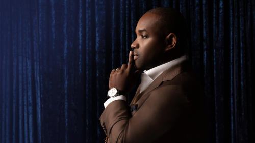 Lawrence Brownlee, a black man in a brown suit, stands in profile against a deep blue backdrop. He raises one finger just under his nose, in front of his lips and chin.