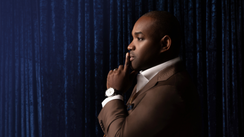 Lawrence Brownlee, a black man in a brown suit, stands in profile against a deep blue backdrop. He raises one finger just under his nose, in front of his lips and chin.
