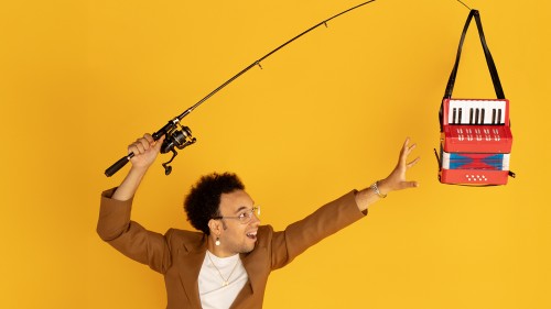 A man in a brown jacket holds a fishing pole over his head and a toy accordion hands from the tip of the fishing pole like a fish.