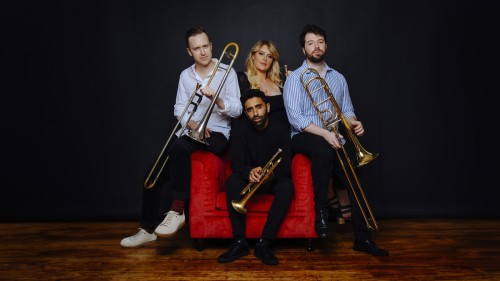 A mixed group of four musicians surround a red armchair; two men sit on the arms of the chair holding trombones in collared shirts, one man sits centered in the chair in a dark outfit holding a trumpet, and behind him a woman with long blond in a black dr