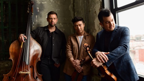 In a shabby industrial looking room, three men hold their stringed instruments.