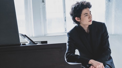 In front of a glassy window, a man with dark, curly hair sits in a black jacket leaning on the lid of the keys on a grand piano. 