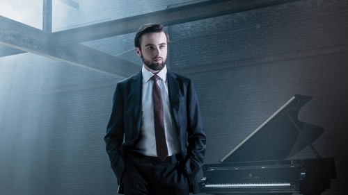 A young bearded white man, under cool light in a blue suit in an industrial building, stands with his hands in his pockets. A grand piano is in the background.