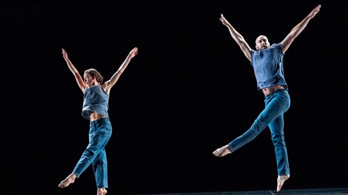 Two dancers leap with arms straight and pointing upwards in a V with one leg pointing down and one pointing a cross their body. 