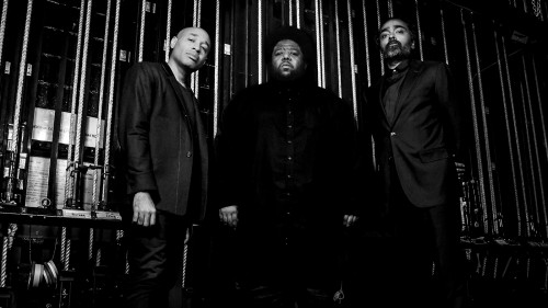 A black and white photo of three stoic black men standing in front of backstage rigging. 