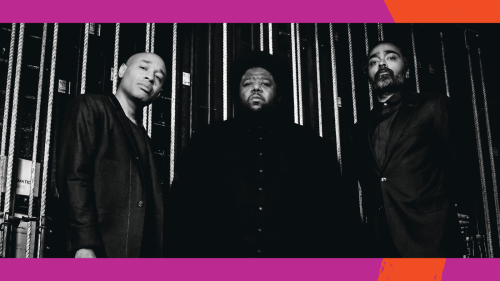 A black and white photo of three stoic men standing in front of backstage rigging. 