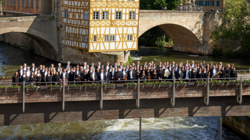The members of the bamberg symphony, on an old stone bridge over a rushing river, in front of an arched bridge that is split by a yellow half-timbered building
