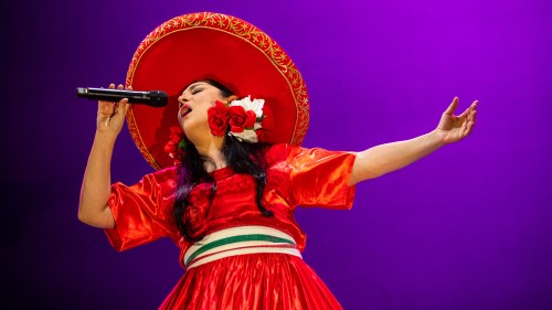 a dark-haired woman sings passionately, one arm extended. she wears a red dress with a yellow belt and broad-brimmed red hat with white and red flowers behind her ear