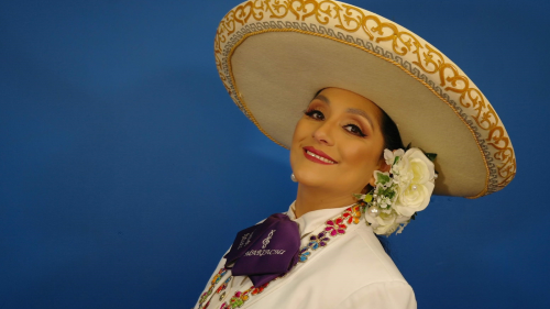 A portrait photo of a smiling woman wearing an elaborate mariachi outfit with a large, tan sombrero and white flowers in her hair. 