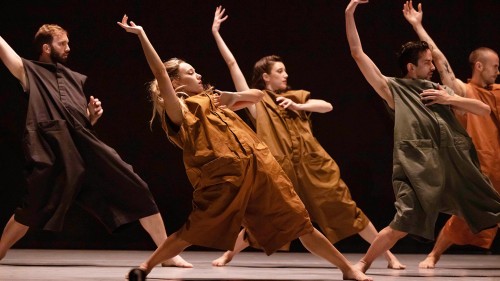 a dance company, men and women wearing baggy rompers in earthy colors. their feet are apart, and they lean back, each with an arm flung above their head.