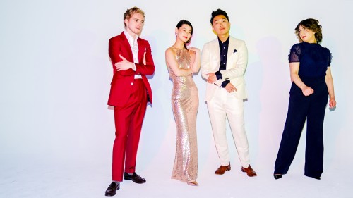 Two men and two women of a string quartet wear bold, elegant outfits, and stand before a bright backdrop under colorful light that casts multiple shadows.