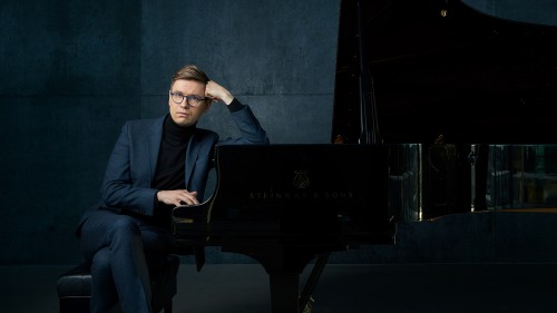 A white man with round glasses in a blue suit in front of a blue background rests an elbow on the top of a piano, his fist at his temple and his other hand idly playing the keys without looking.