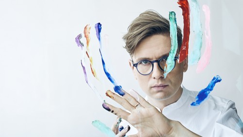 A white man with small round glasses stares intensely ahead while using finger paint on a plexiglass panel in front of his face. 