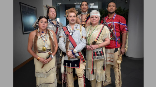 A mixed group of six Indigenous performers in traditional dress stand together with gentle smiles in a white hallway. 