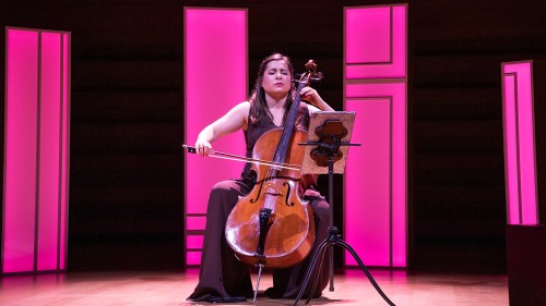 A white woman plays a cello on a stage lit in pink, her brow furrowed as she performs passionately