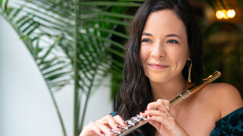 a woman with long dark hair and a black top plays the flute outdoors in front of green trees