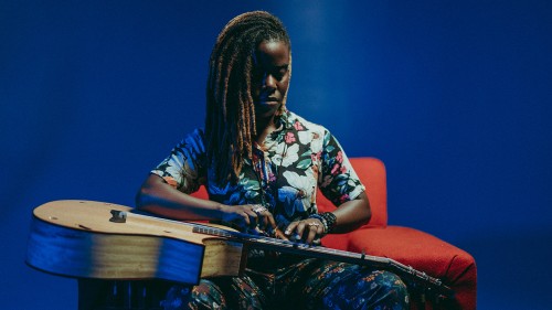 Yasmin Williams, a black woman in a multicolored top, sits on a red chair in front of a blue background playing a guitar sitting on her lap horizontally. 