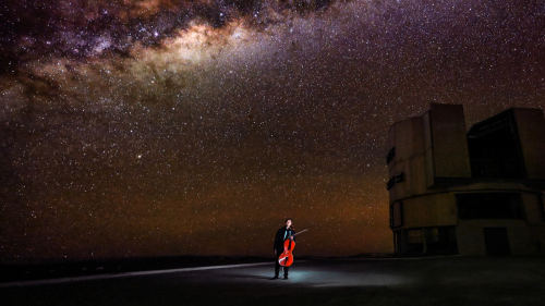 Yo-Yo Ma, under a small white spotlight in the Atacama Desert. The galaxy spans the dark sky above him, and a large rock outcrop looms at his left, a dark space in the bright sky..