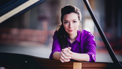 A white woman with pulled-back long brown hair wearing a bright purple shirt faces the camera as she leans over the keys of a grand piano, her face framed by the lid and the stick of the piano.  