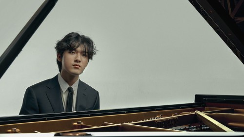 A young, South Korean man sits at the piano, his face framed by the stick of the lid, staring solemnly ahead in a dark suit.