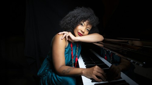 A Cuban woman with dark curly hair leans her head on her arm on the piano while another hand lays on the piano keys. 