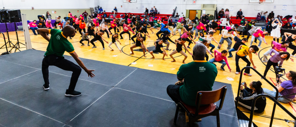 facing away, a black man in a green t-shirt leads a class of 150 people in movements from Revelations. Everyone's feet are wide, their knees bend out, and they reach down and to their right with one hand.