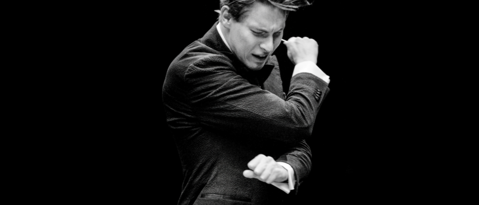 a black and white image of a white man in his 20s conducting energetically, his arms crossed diagonally across his chest