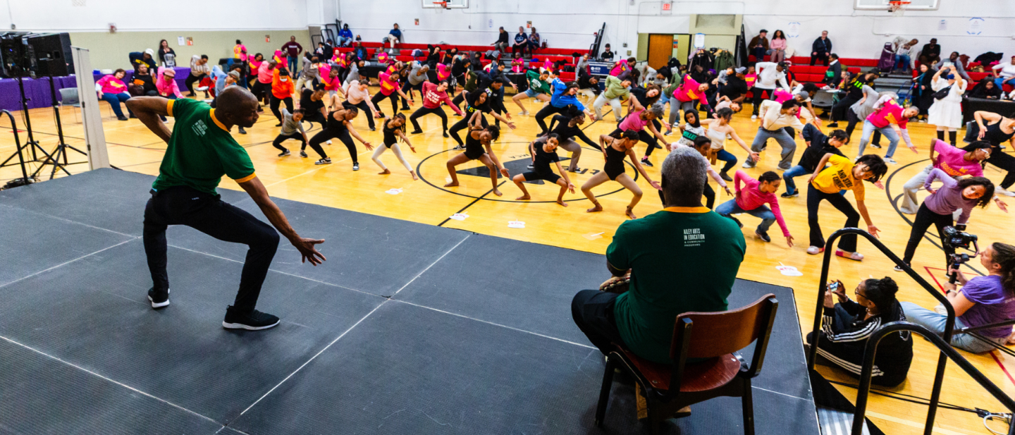facing away, a black man in a green t-shirt leads a class of 150 people in movements from Revelations. Everyone's feet are wide, their knees bend out, and they reach down and to their right with one hand.