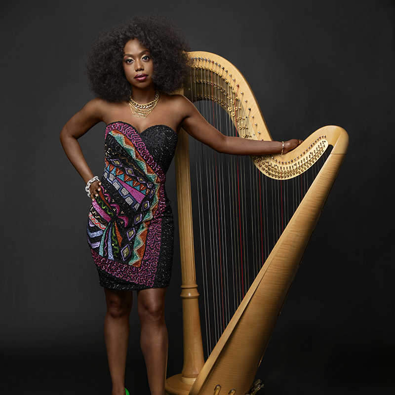A Black woman poses with a golden harp in front of a dark background in a short, strapless dress with a multi-colored geometric pattern.