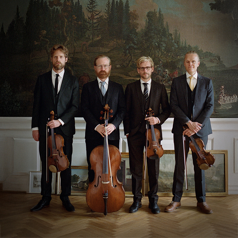 Four white men in a string quartet stand in a row in dark suits holding their instruments in front of several nature murals. 