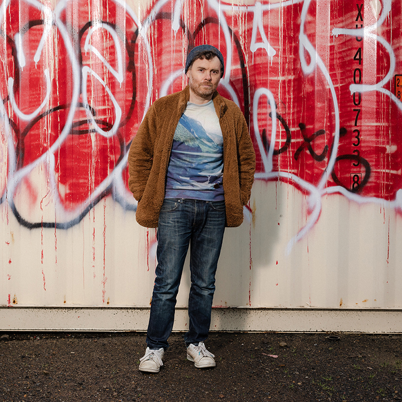 A white man stands in blue jeans, a t-shirt with mountains, and a brown fuzzy jacket in front of a wall of graffiti. 