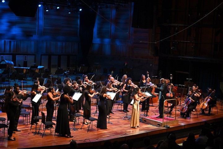 24 musicians from Chineke! Orchestra in black outfits stand on stage, playing their instruments. A woman in a gold dress stands front center, playing a violin.