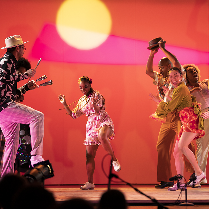A multicultural group of five dancers clap and dance joyfully in front of a pink and orange background. 