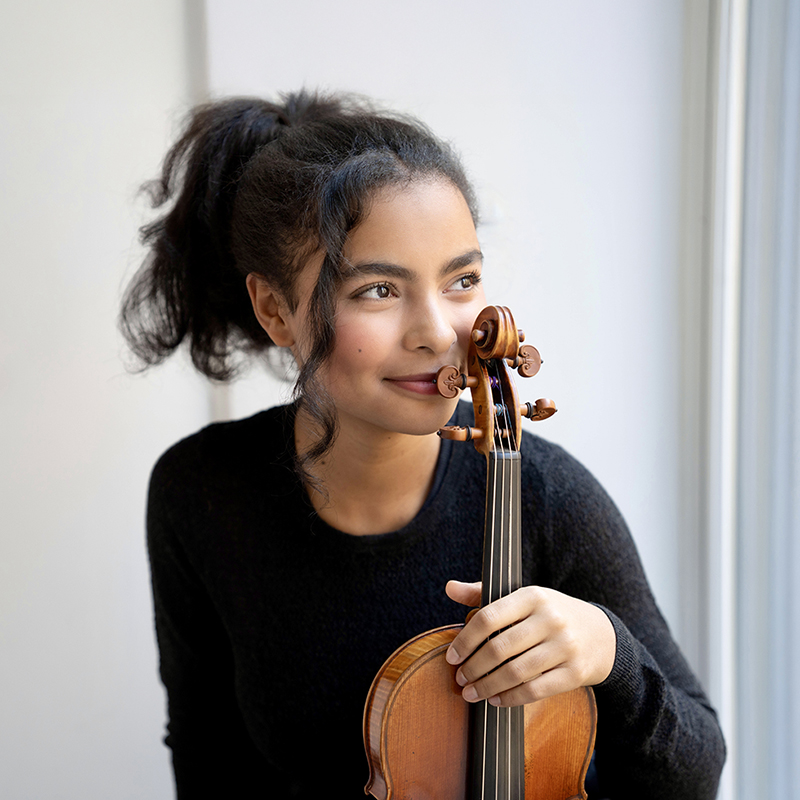 A young woman holds her violin upright wearing a black sweater with her dark curly hair pulled back in a ponytail.