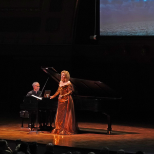 a man plays a grand piano and a white woman in a copper gown performs in front of a screen with an image of the sky