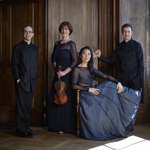 Four members of a string quartet, two men flanking two women, are inside an ancient mansion. One of the women holds a violin. 
