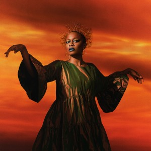 in front of an apocalyptic sunset backdrop, a black woman stands with arms elegantly outspread. her head is wreathed in wires and her robe and styling is coppery and metallic
