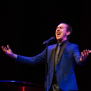 a white man in a blue jacket sings, his eyes closed and arms outspread