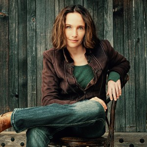 a brown haired white woman in a brown suede jacket and jeans crosses one leg over the other