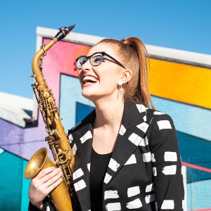 a young white woman smiles as she holds her saxophone in front of a vivid mural