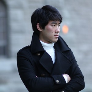 a young chinese-canadian man folds his arms as he stands outdoors in a dark wool coat and white turtleneck