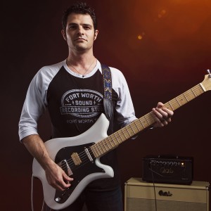 a white man in a black and white t-shirt holds a guitar