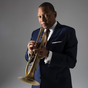a black man holds his trumpet almost to his mouth, he wears a dark gray suit and is in a photo studio with a gray background