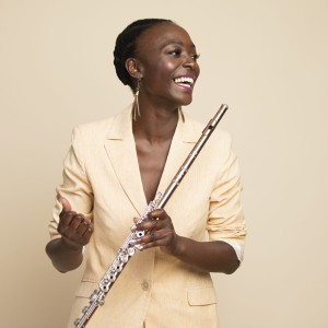 a smiling black woman in a cream-colored suit holds her flute and looks to the side