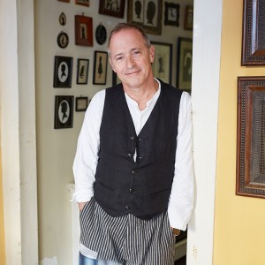 A white man in his mid-60s wears a loose vest and slouchy trousers leans against a doorway. 